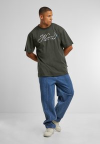 Dark green cotton t-shirt with oversized fit, featuring a shiny, multicolored "Hand" graphic. Paired with light blue, relaxed-fit jeans.