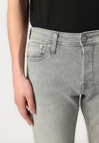 Grå denimbukser med en tettsittende design, synlige framlommer og metallbeslag. Stoffet har en jevn tekstur med subtile sømdetaljer.