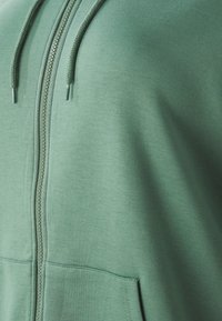 Zelená mikina na zips vyrobená z mäkkej látky, ktorá má hladkú textúru, šnúrky a klokanie vrecko vpredu.