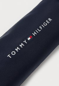 Mörkblå, strukturerad tygväska med vit "TOMMY HILFIGER"-logotyp och röda, vita och blå accenter. Kompakt cylindrisk design.