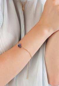 Gouden kettingarmband met een cluster van blauwe stenen, tegen een gladde huid. Fijne, verstelbare ketting met een klein sluitingdetail.