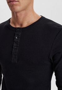 Schwarzes Langarm-Henley-Shirt aus geripptem Baumwollstoff, mit Rundhalsausschnitt und einer Knopfleiste mit drei Knöpfen am Kragen. Texturiertes Material.