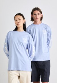 Light blue long-sleeve t-shirts with white floral and tentacle designs on sleeves. Model wearing cream and dark shorts. Cotton material.