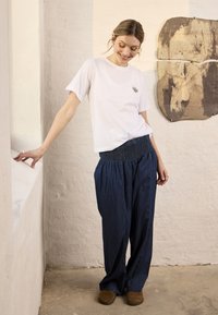Femme portant un t-shirt blanc et un pantalon ample bleu marine, debout à l'intérieur, souriante et appuyée sur un rebord blanc avec une œuvre murale abstraite derrière elle.