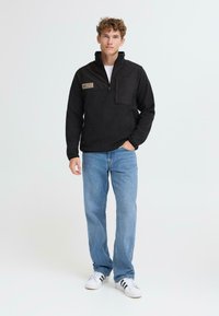 Sudadera de poliéster negro con media cremallera, que presenta un bolsillo en el pecho y un parche de tela. Combinada con jeans de mezclilla azul claro y zapatillas blancas.