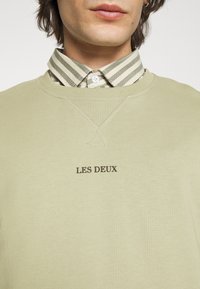 Jasnozielona bluza z prążkowanej tkaniny, z okrągłym dekoltem i nadrukowanym logo "LES DEUX" w ciemnym brązie. Nałożona na koszulę w paski.