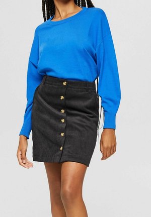 Mujer con una blusa azul brillante de manga larga metida dentro de una falda de pana negra con botones en la parte delantera, de pie frente a un fondo liso.