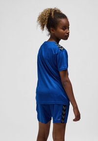 Blå atletisk tröja och shortsset av lättviktigt tyg, med svarta chevrondetaljer på axlar och shorts. Mjukt textur.