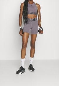 Szary sportowy krótki top i szorty z wysokim stanem z czarnymi paskami akcentującymi, w połączeniu z czarnymi trampkami i białymi skarpetkami. W ręku trzyma opaskę do ćwiczeń.