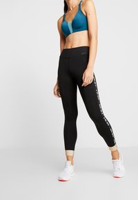 Žena v modrém sportovním topu, černých legínách Nike s nápisem "Just Do It" na boku a bílých sportovních botách, stojí na bílém pozadí.