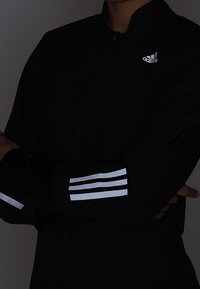 Veste de sport noire avec un col haut, dotée d'accents argentés à trois rayures sur les manches et d'un petit logo blanc sur la poitrine.