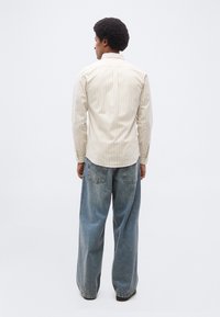 Personne aux cheveux courts et bouclés portant une chemise à rayures beige et blanche boutonnée et un jean large bleu, debout et de dos sur un fond blanc.