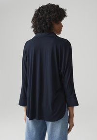 Blusa leggera di colore blu navy, con colletto, maniche lunghe e vestibilità comoda, abbinata a jeans in denim chiaro. Texture liscia.