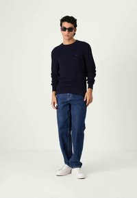 Maglione a coste navy, jeans blu dal taglio rilassato e sneakers bianche. Accessorio: occhiali da sole scuri. Un design minimalista con un sottile dettaglio di branding.