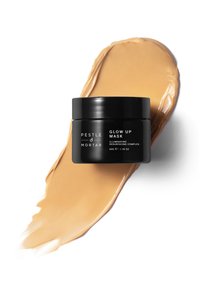 Pestle & Mortar Glow Up Maske in einem schwarzen Glas mit weißem Text. Eine cremige beige Maske ist neben dem Glas aufgetragen, um eine glatte Textur zu zeigen.