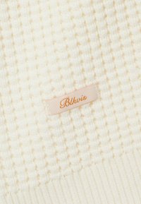 Tissu tricoté texturé de couleur crème avec une petite étiquette beige cousue, portant un texte cursif orange disant "Blkvis".