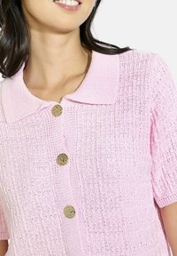 Gaiši rozā adīta blūze ar īsām piedurknēm, apkakles galu un trim dekoratīvām zeltītām pogām. Teksturēts audums ar režģa rakstu.