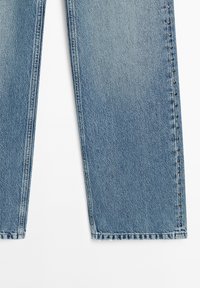 Světle modré džíny z denimu se zdviženými nohavicemi, s neupraveným lemem a malými stříbrnými cvočky podél bočních švů. Měkká textura a klasický design.