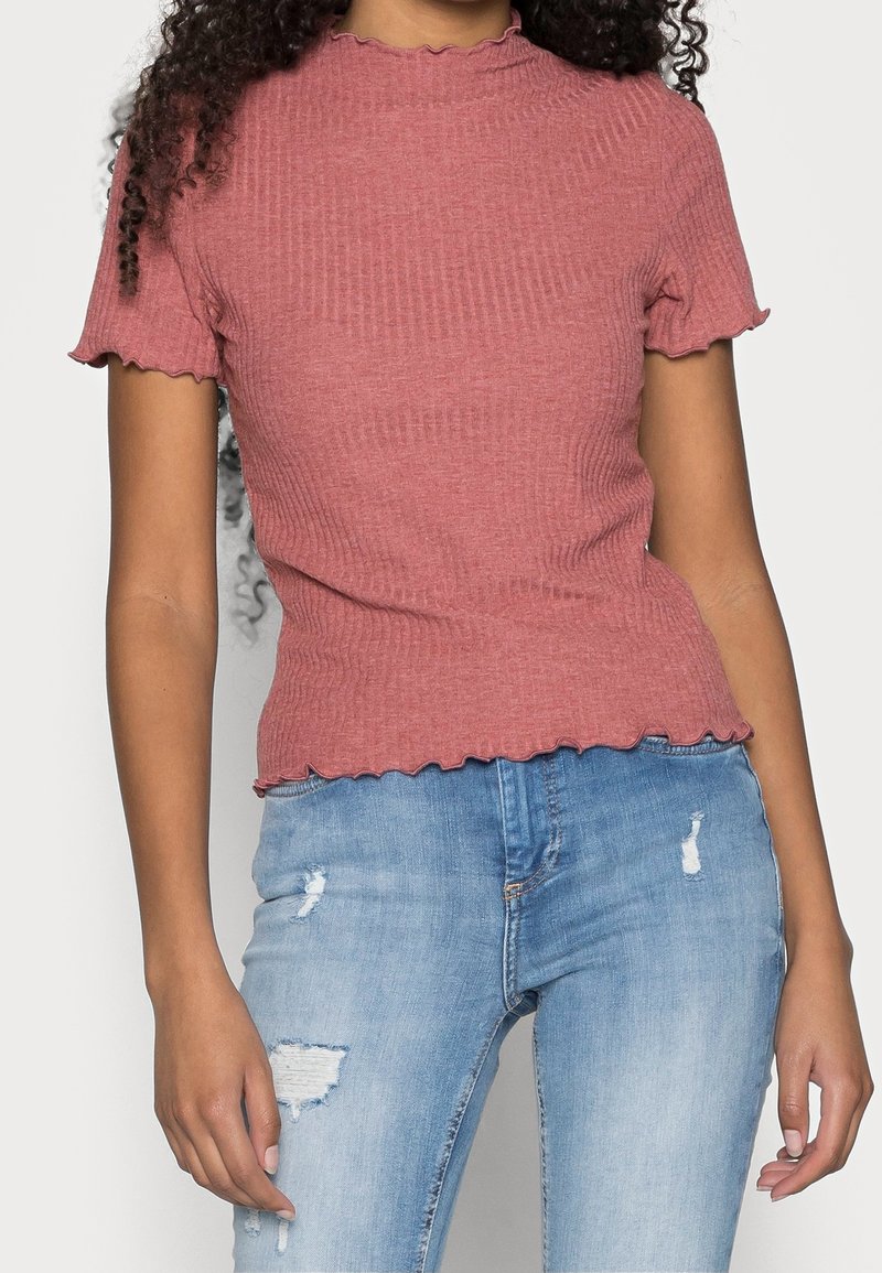 Haut à manches courtes côtelé en rose poussiéreux avec un col haut à bords festonnés et un ourlet effiloché. Associé à un jeans déchiré bleu clair.