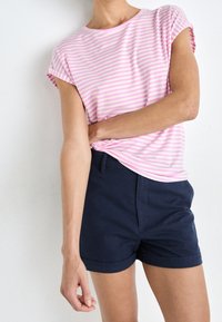 En person som bär en kortärmad skjorta med rosa och vita ränder och hög midja på navyblå shorts, poserar mot en enfärgad vit bakgrund.