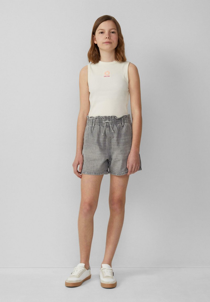 Kremowy bezrękawnik z małą grafiką, zestawiony z szarymi szortami dżinsowymi z elastycznym paskiem. Białe sneakersy z beżową podeszwą.