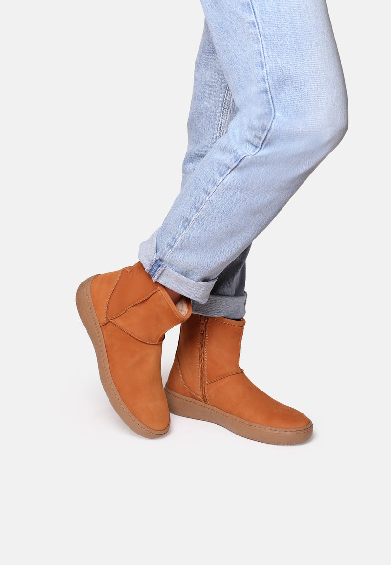 Bottines beige en suède avec fermeture éclair sur le côté et semelle en caoutchouc. Portées avec un jean léger bleu retroussé, mettant en avant un look décontracté.