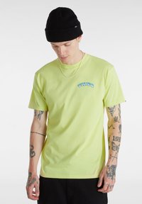 Limevihreä puuvillapaita lyhyillä hihoilla, jossa on sininen "VANS" -logo rinnassa. Musta pipo ja näkyvät tatuointiyksityiskohdat käsivarsilla.