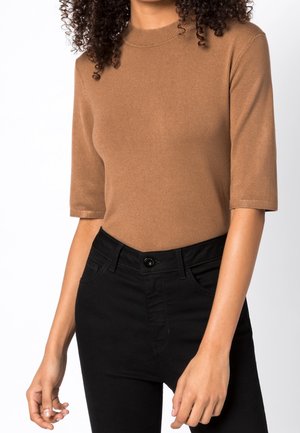Femme portant un haut en maille marron à manches courtes associé à un pantalon noir taille haute, debout les bras détendus le long du corps.