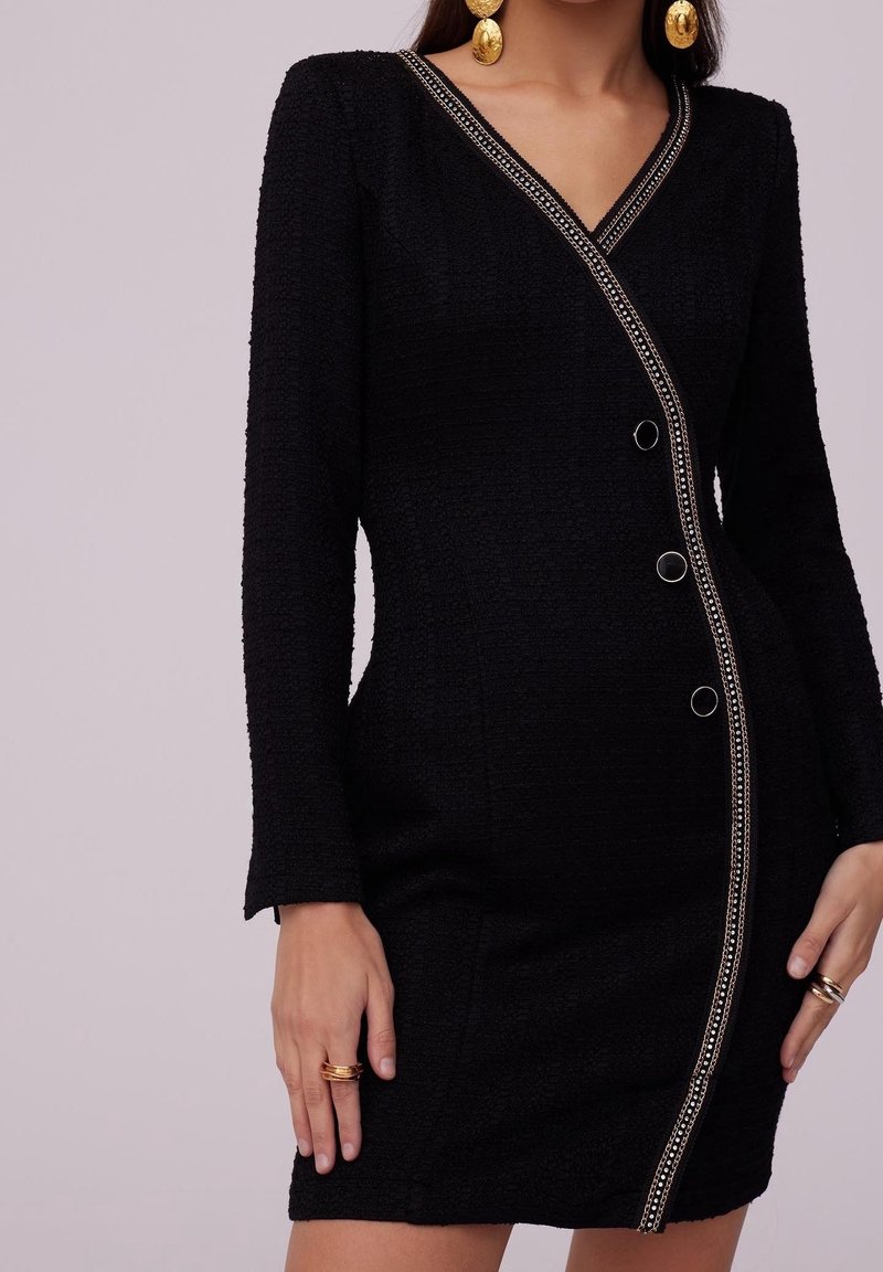 Femme portant une robe noire texturée avec des boutons diagonaux ornés de chaînes, associée à de grandes boucles d'oreilles dorées et des bagues en or.