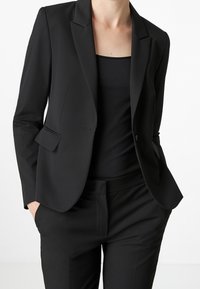 Černé sako na míru s klopovým límcem, předními kapsami a elegantním designem. Nahoře je nošeno přes černý top s hladkou texturou.