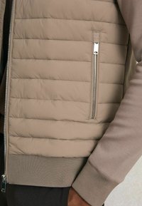 Beige quiltet vest med en glat finish, sidelukning og ribbet kant. Blødt stof og neutrale farver skaber et let og afslappet design.