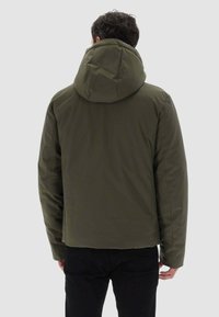 Uomo che indossa una giacca a vento verde oliva con cappuccio, visto da dietro, in piedi contro uno sfondo grigio chiaro uniforme.