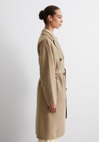 Cappotto in lana beige a doppio petto con maniche inserite, colletto a revers e vita con cintura, mostrato su un modello in vista laterale.