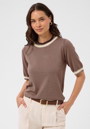 Femme aux cheveux bruns ondulés portant un haut marron à manches courtes, un pantalon crème, des boucles d’oreilles dorées, un collier foncé à perles et les mains dans les poches.