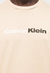 Εγγύς πλάνο μιας μπεζ μπλούζας με το λογότυπο «Calvin Klein» τυπωμένο στο στήθος με λευκά και μαύρα γράμματα.