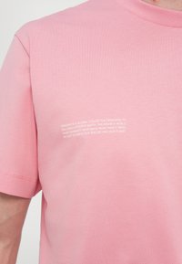 T-shirt in cotone rosa chiaro con maniche corte e scollatura a girocollo. Il testo bianco sul lato sinistro parla della missione di sostenibilità del brand.
