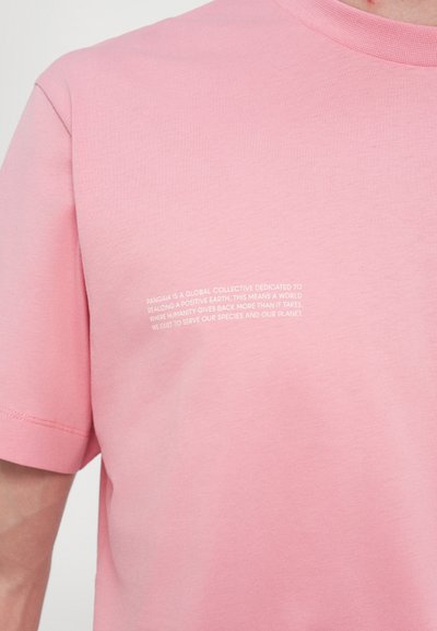 T-shirt en coton rose clair avec des manches courtes et un col rond. Un texte blanc sur le côté gauche évoque la mission de durabilité de la marque.