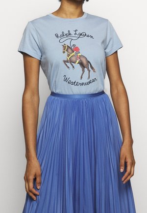 Woman wearing a light blue Ralph Lauren Westernwear t-shirt with a horse and rider graphic, paired with a high-waisted blue pleated skirt.