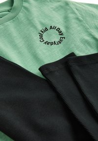 Grünes Baumwollhemd mit schwarzen Rippärmeln, mit dem kreisförmigen Text "Cool Kid All Day Every Day" in Schwarz im Brustbereich.