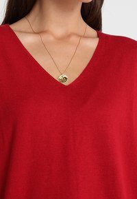 Maglione rosso a V in tessuto morbido, con una texture liscia. Una collana con pendente dorato poggia sul collo.
