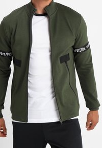 Giacca zip-up verde oliva realizzata in tessuto morbido, con dettagli neri sulle maniche con testo, colletto alto e design aderente.
