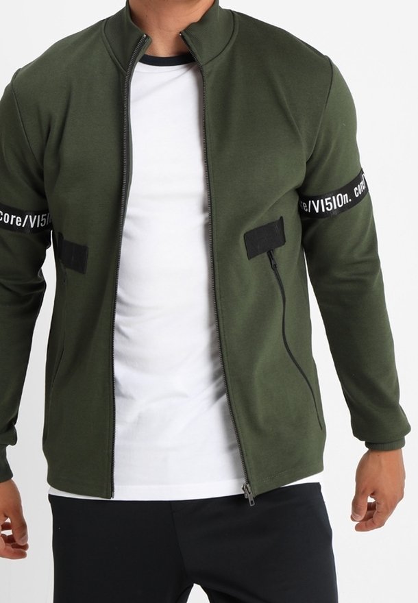 Giacca zip-up verde oliva realizzata in tessuto morbido, con dettagli neri sulle maniche con testo, colletto alto e design aderente.