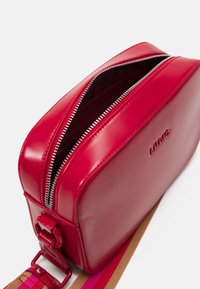 Red faux leather crossbody bag with a double zipper, adjustable striped fabric strap, and embossed logo on the side.