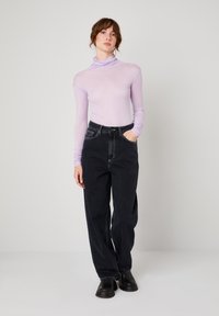 Žena stoji s jednom rukom u džepu, noseći svijetloljubičasti rol-krojek, široke crne traperice i crne čizme na jednostavnoj pozadini.