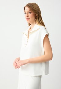 Witte rits-top met een kraag, korte mouwen en een gouden ritsaccent. Glad, lichtgewicht materiaal met een ontspannen pasvorm en geen zichtbare patronen.
