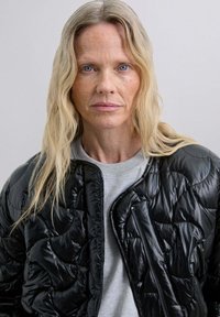 Chaqueta acolchada negra con una textura brillante y quiltada, que presenta un bolsillo con cremallera y un escote redondeado, superpuesta sobre un sudadera gris.