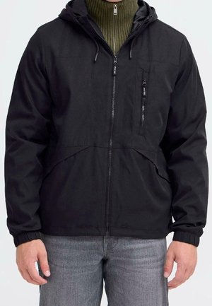 Homme portant une veste noire à capuche par-dessus un pull vert zippé et un jean gris, debout les bras détendus le long du corps.