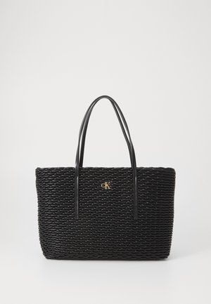 Svart vevd tote bag med teksturert overflate, med forlengede svarte håndtak og en gull "CK"-logo midt foran.