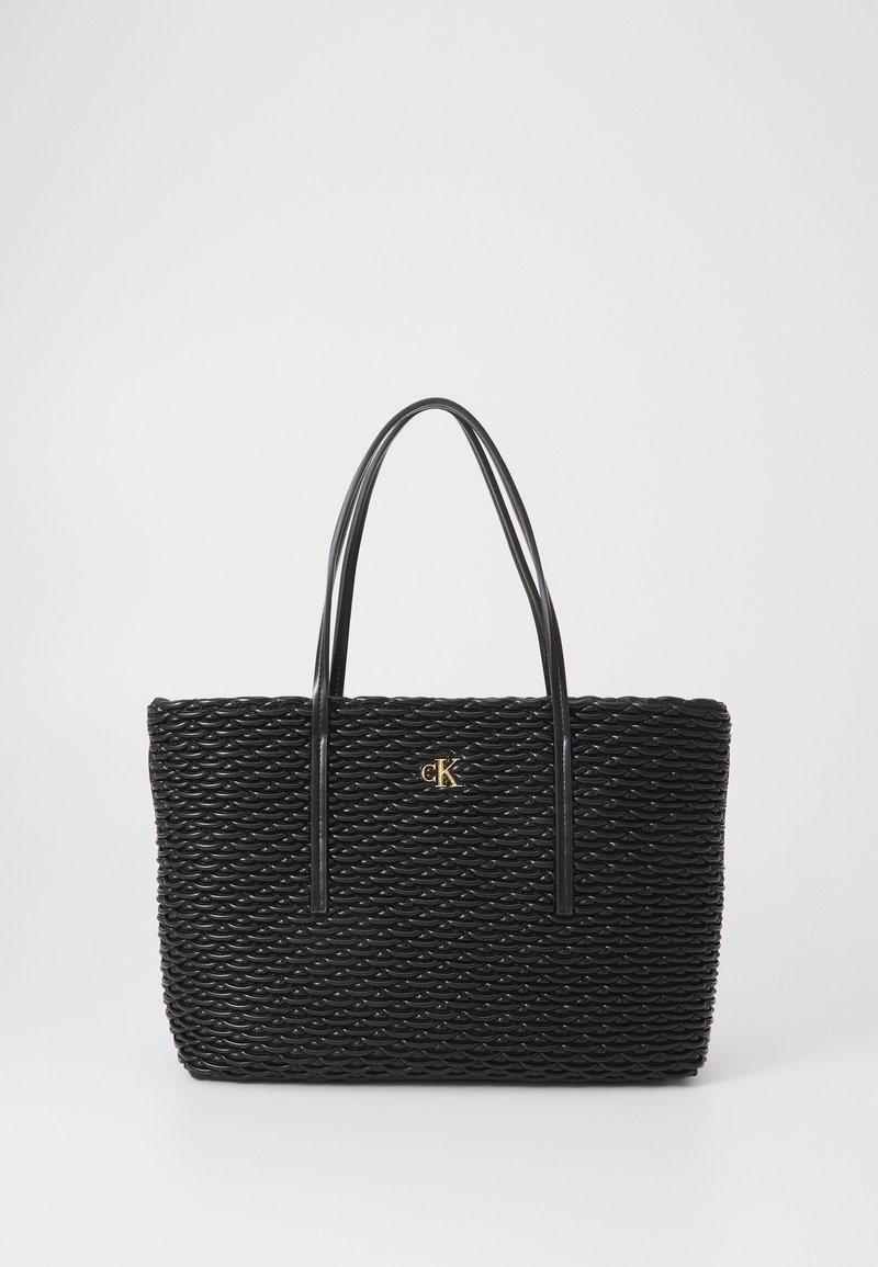 Svart vevd tote bag med teksturert overflate, med forlengede svarte håndtak og en gull "CK"-logo midt foran.