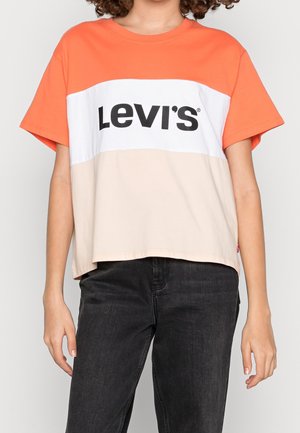 Personne portant un T-shirt à manches courtes en blocs de couleurs orange, blanc avec le logo "Levi's" et beige, associé à un jean noir.
